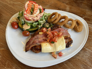 A delicious plate featuring grilled chicken topped with bacon, cheese, salsa, alongside a fresh salad and crispy onion rings.