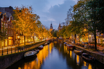 As twilight descends on Amsterdam, the charming canals reflect the golden hues of autumn trees. Shops glow warmly while boats float serenely, creating a magical atmosphere in this historic city.