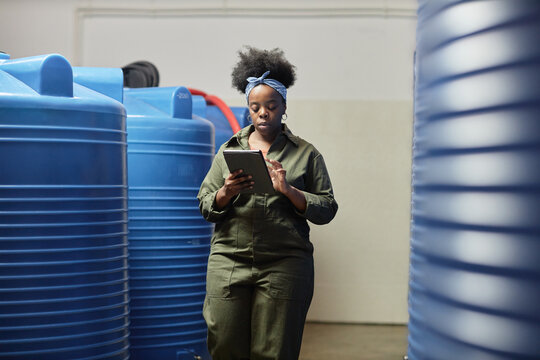 Medium full shot of female African American process technician writing on tablet computer walking along huge plastic barrels while inspecting apple maceration at craft cider factory, copy space - Powered by Adobe