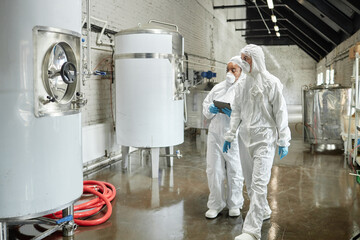 Full length shot of professional female supervisor in mask and coverall walking towards stainless tank with liquid while inspecting workshop equipment with colleague at factory