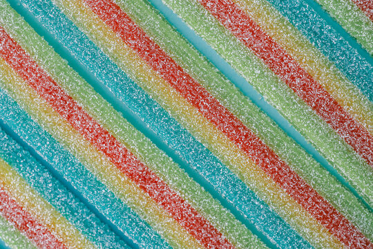 Colorful sugar-coated rainbow candy strips laid in parallel rows on a light blue background. The vibrant colors and texture highlight the sugary details, making it perfect for sweet and festive themes