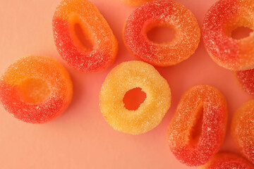 Close-up of sugar-coated gummy candy rings in vibrant red, orange, and yellow tones placed on a soft peach-colored background. The sugary texture and bright colors create a playful and sweet visual