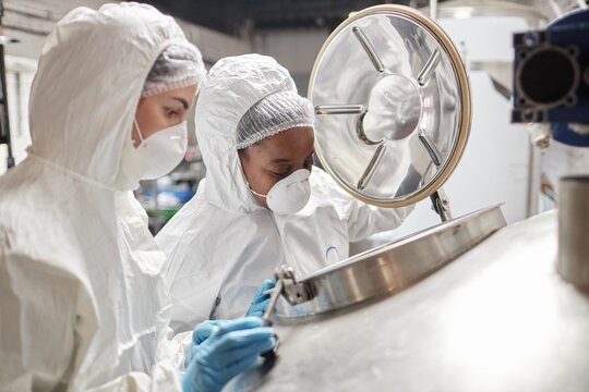 Medium shot of African American female process technician in mask opening tank lid while inspecting product fermentation with colleague in factory workshop, copy space
