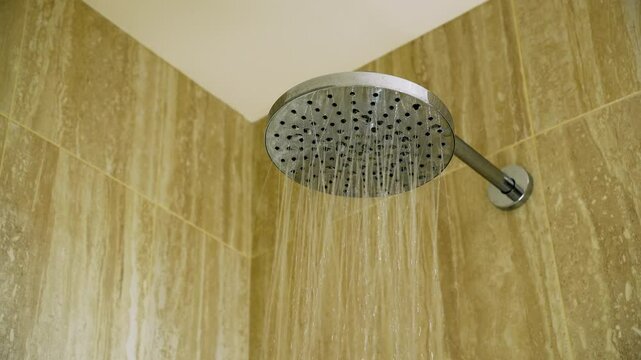Modern rainfall shower head with running water against beige bathroom wall. Chrome shower head spraying fresh water streams showing the details of the water droplets slow motion.