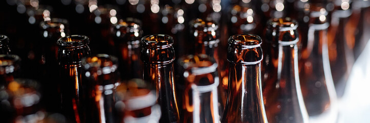 Header image of dozen open glass bottles shining in light at production line in craft beer factory,...