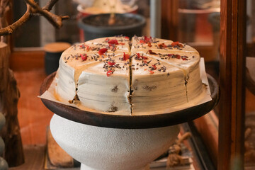 Close up of delicious cream lavender cake for sale in a glass showcase in cafe bakery. The sliced whole cake is decorated with flower petals. Pretty, warm rustic mood.