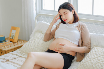 Young Asian tried pregnant woman have a morning sickness is sitting on bed and touching her forehead. pregnancy, motherhood, people and expectation concept