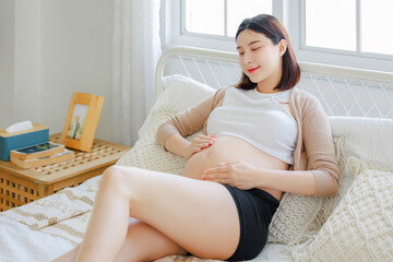 Young Asian happy pregnant woman is sitting and relaxing on bed and and touching her belly. Family plan, pregnancy, motherhood, people and expectation concept
