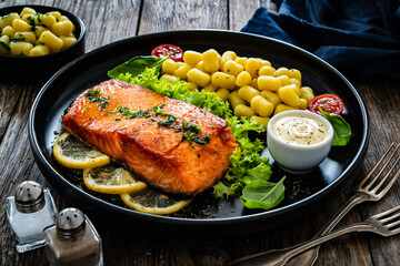 Seared salmon steak with gnocchi and lemon slices served on black plate on wooden table
