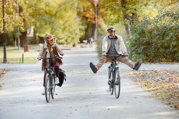 Naklejka premium joyful senior couple riding bicycles in an autumn park, with the man playfully raising his legs in the air, enjoying an active and carefree moment outdoors.