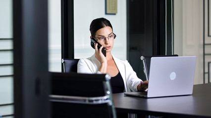 Confident businesswoman working on laptop and talking on phone in office. The concept of business negotiations, consultation and effective work in a modern business environment.