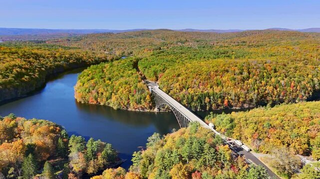 Aerial footage of French King Bridge, Massachusetts, and Connecticut River surrounded by a forest of trees in vibrant fall foliage basking in warm sunlight under blue skies.