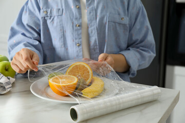 Woman putting plastic food wrap over plate with fruits at countertop in kitchen, closeup