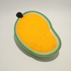 The image shows a sponge shaped like a mango with a green border and a small black stem. This is interesting due to its unique and playful design, resembling a fruit.