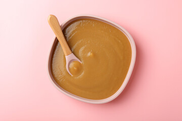 Delicious baby food with spoon in bowl on pink table, top view