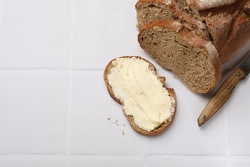 Fresh bread with butter and knife on white tiled table, flat lay. Space for text