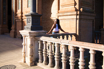 Fototapeta premium young and pretty brunette Moroccan woman dressed in white top and jeans is happy leaning on the railing in the square of Spain in Seville, she looks back and takes the photo.