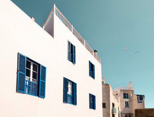 white buildings with blue shutters in essaouira medina near the port, morocco, with a bright blue sky and seagulls flying above, showcasing traditional architecture on a sunny day in the historic city