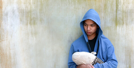 Asian teenboy in blue hoodie holding clothes doll and standing in front of dirty wall, look sad,...