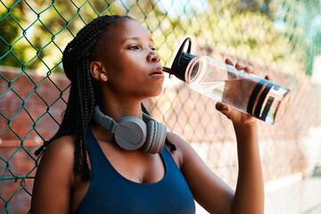 Fitness, court and black woman with water for drinking, hydration and break for basketball competition. Fence, female person and athlete with rest for liquid beverage, sports game and h2o for thirst