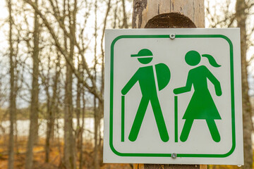 Green hiking trail sign featuring icons of a man and woman with walking sticks. The sign is mounted on a wooden post, with a blurred forest backdrop and bare trees. - Powered by Adobe