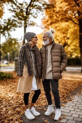A couple of older people are standing in the fall, smiling at the camera. They are wearing warm clothing and appear to be enjoying the season
