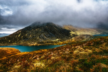 A scenic view of a mountain range, with a foreground of dry grass, a curved lake in the valley, and a hilltop shrouded in mist.