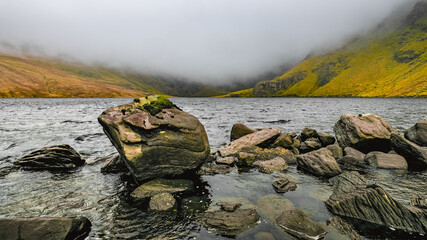 A lake stretches out in front of a misty hillside. Large, dark rocks protrude from the water near...