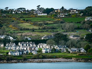 A view of a small town nestled in a green, rolling hillside. There are trees scattered throughout the landscape, and a body of water is visible in the background.