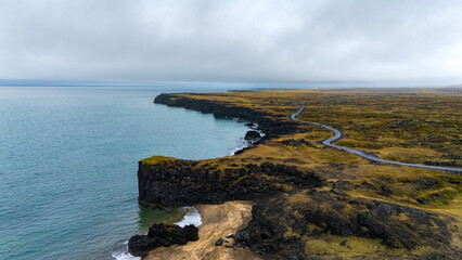 A breathtaking aerial view of a coastal mountain landscape. Steep, rocky cliffs rise sharply from the water, meeting with a winding road that skirts the shoreline