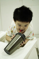 Asian boy drinking water in silver glass, sitting and smiling on high chair at home