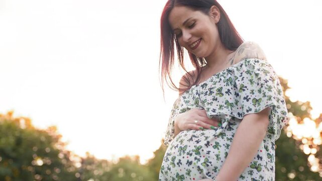 Pregnant caucasian woman in summer dress posing outdoors. Close up of pregnant caucasian woman in summer dress posing outdoors. Attractive brunette caressing her tummy while standing at green garden.