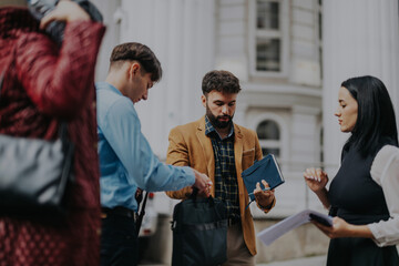 Obraz premium A group of business people engaged in a conversation outdoors in the city, holding documents and a tablet. The setting is urban, emphasizing collaboration and teamwork in a professional environment.