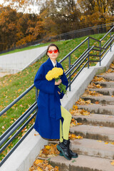 Portrait of stylish smiling young woman walking in park, wearing blue coat. Autumn trendy fashion