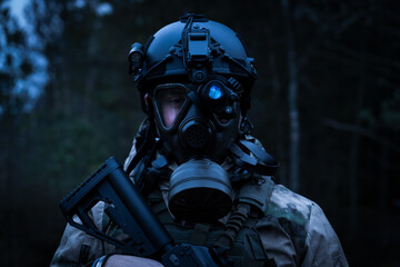 A military soldier in a gas mask and a helmet with a night vision device in the forest at dusk.