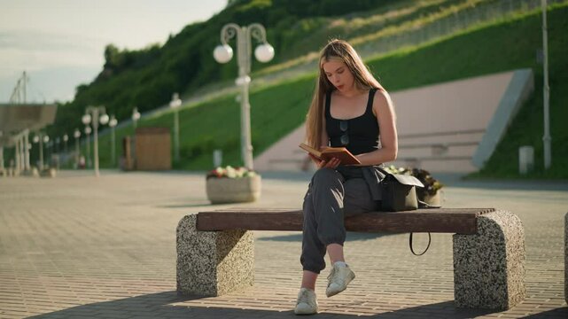 Woman seated outdoors on a bench with legs crossed, flipping a page from her book and observing a piece of paper thoughtfully, the background features lamp poles and a person seated in the distance