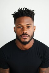 A man with short dreadlocks, wearing a black shirt, gazes confidently against a plain background, exuding modern style, assurance, and depth in expression.