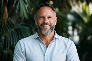Fototapeta premium A joyful portrait of a middle-aged man with a beard and a warm smile, dressed in a light-colored shirt, standing against a lush garden background full of greenery.