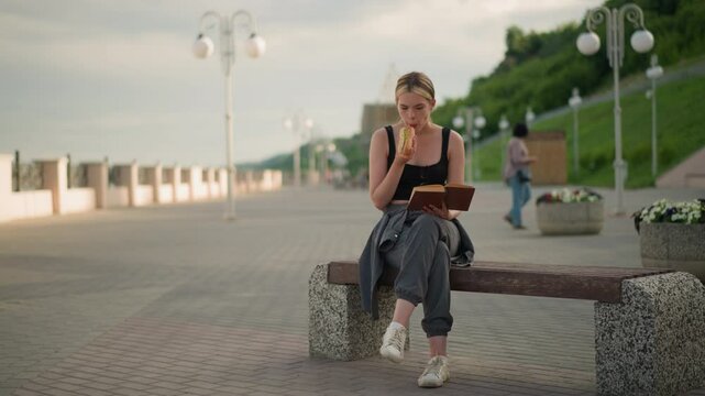 Woman seated outdoors with crossed legs, holding a book in one hand while eating snacks and reading, the blurred background features a lamp post lined up with a blur view of a woman walking
