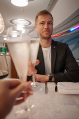 A man sits at a dining table in a stylish restaurant, wearing a dark blazer and white shirt. A hand holds a champagne flute, with sleek design elements in the background.