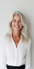 Fototapeta premium A cheerful woman with gray hair beams in a crisp white blouse, radiating positivity and confidence in front of a bright, minimalist background.