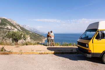 Couple enjoying the view of Croatian mountainous coast standing by their retro yellow camper van parked on a cliff, Croatia