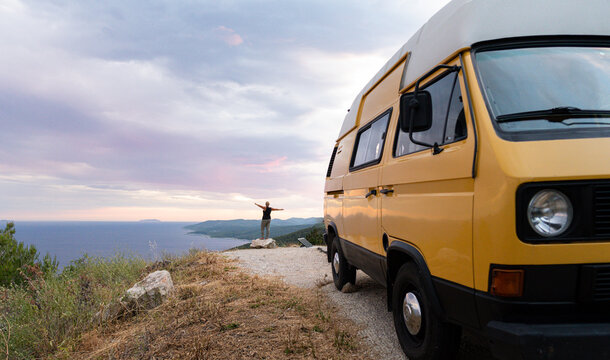 Yellow retro camper van parked on a dirt road above the sea on Korcula island, Croatia. Small female silhouette standing on the cliff with her hands up.