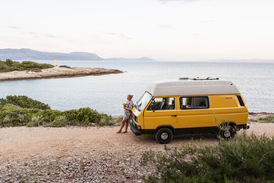 Young blonde woman enjoying the sea view in her yellow retro camper van parked on a dirt road above the sea on Korcula island, Croatia