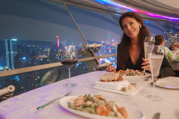 A woman enjoys a meal at an elegant restaurant overlooking Shanghai's skyline, featuring the illuminated Oriental Pearl Tower and modern architecture.