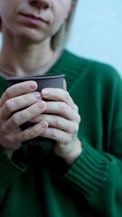 hands of a sick woman in a green sweater holding a cup with a hot drink
