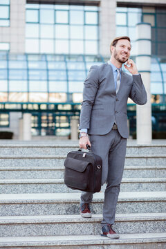 Young Caucasian businessman walking down the stairs in front of an office building. Carrying a briefcase and talking on the phone