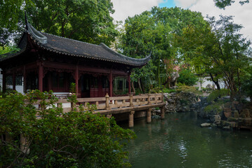 A traditional Chinese pavilion with curved eaves and wooden detailing stands by a pond. Lush greenery surrounds the scene, reflecting in the still water.