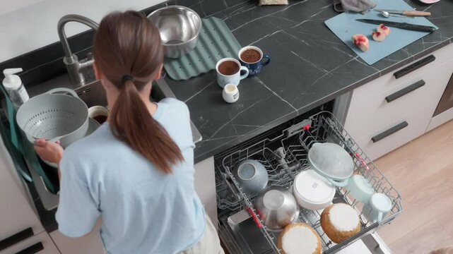 A young woman cleans the kitchen and puts dirty dishes in the dishwasher. A female housewife does the housework and tidies up the kitchen. Daily women's duties and house cleaning.