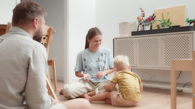 Young parents have fun and carefree playing and messing with their little child sitting on the floor in the room. Mom, dad and son frolic together at home. Pleasant leisure and family time.